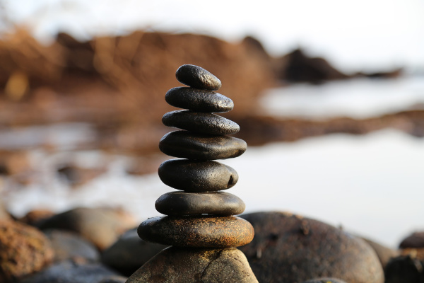 A stack of smooth, dark stones balanced on top of each other outdoors near water, with blurred rocks and water in the background.