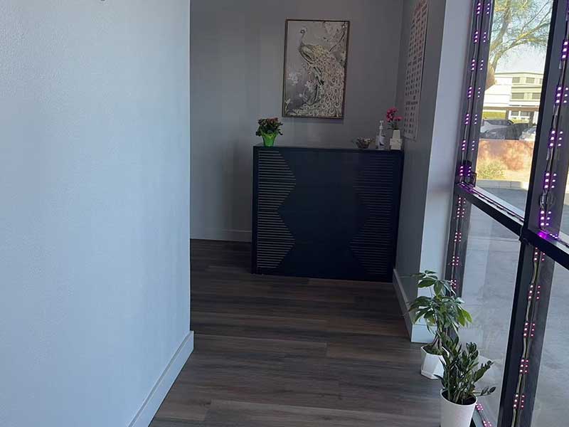 A small reception area with a dark desk, framed artwork above, potted plants, and large windows with purple lights visible on the right.