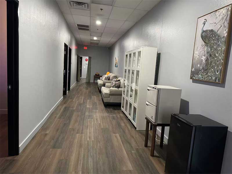 A hallway with wood flooring, gray walls, a sofa, shelving units, a mini fridge, and a peacock painting on the wall. Doors and an exit sign are visible at the end of the hallway.