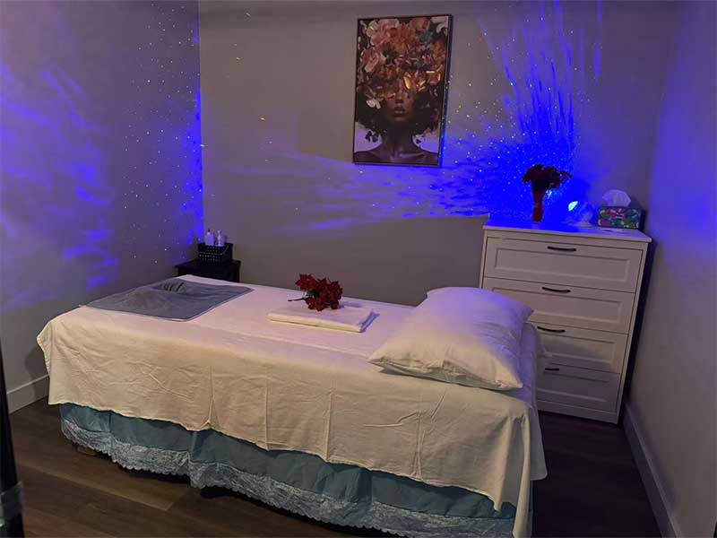 A massage therapy room with a bed, dresser, towel, flowers, tissues, and blue accent lighting on the wall. A floral artwork hangs above the dresser.