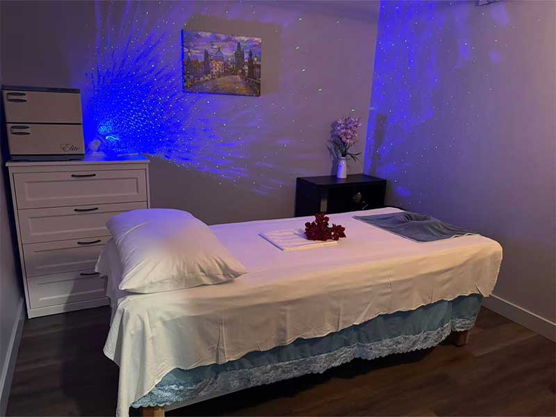 A neatly made massage table in a dimly lit room with blue ambient lights, a dresser, a small table with a vase, and a cityscape painting on the wall.