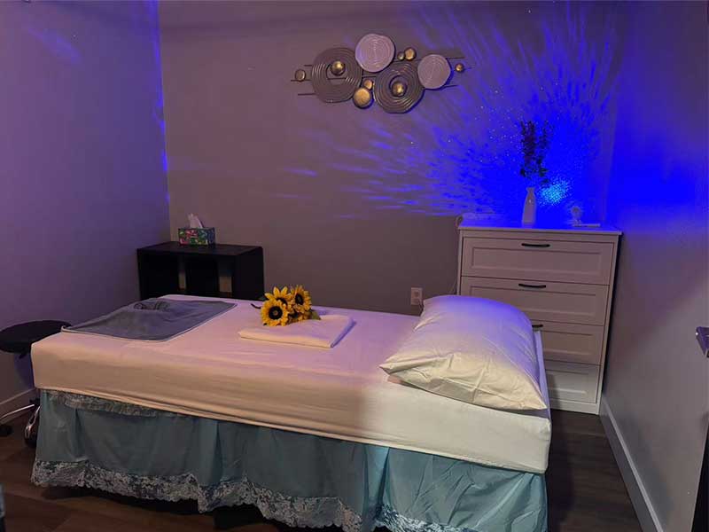 A massage room with a neatly made bed, a towel and sunflowers on top, white dresser, stool, and modern wall art, lit by soft blue lighting.