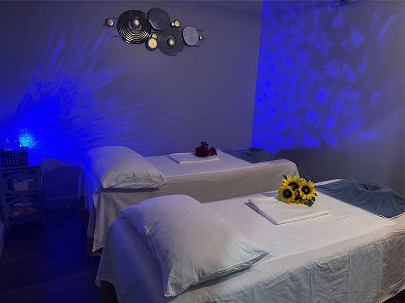 A dimly lit massage room with two beds topped with white linens, one with sunflowers, and abstract wall art; blue ambient lighting enhances the relaxing atmosphere.