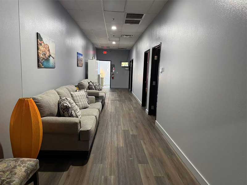 A hallway with wood flooring, gray walls, a sofa with cushions, a chair, wall art, and several doors leading to other rooms. Ceiling lights and an exit door are visible at the end.