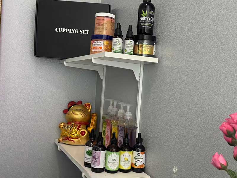 Two white wall shelves hold various wellness products, bottles, dispensers, a black cupping set box, and a gold waving cat figurine against a gray wall.
