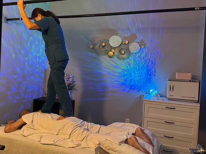A massage therapist stands on parallel bars performing a barefoot massage on a client lying face down on a table in a softly lit room.
