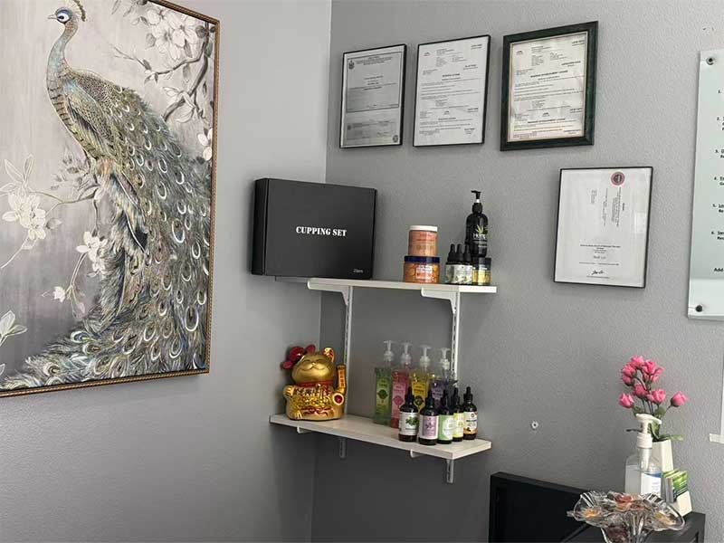 A corner with certificates on the wall, a painting of a peacock, shelves holding skincare products, a cupping set box, a golden cat figurine, and a vase with pink flowers.